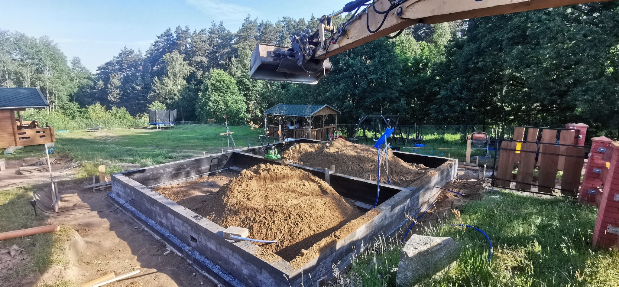 Wypełnianie piaskiem wykopu pod fundamenty domu jednorodzinnego przez koparkę, widoczne szalunki i przygotowany teren działki z domkiem letniskowym w tle.