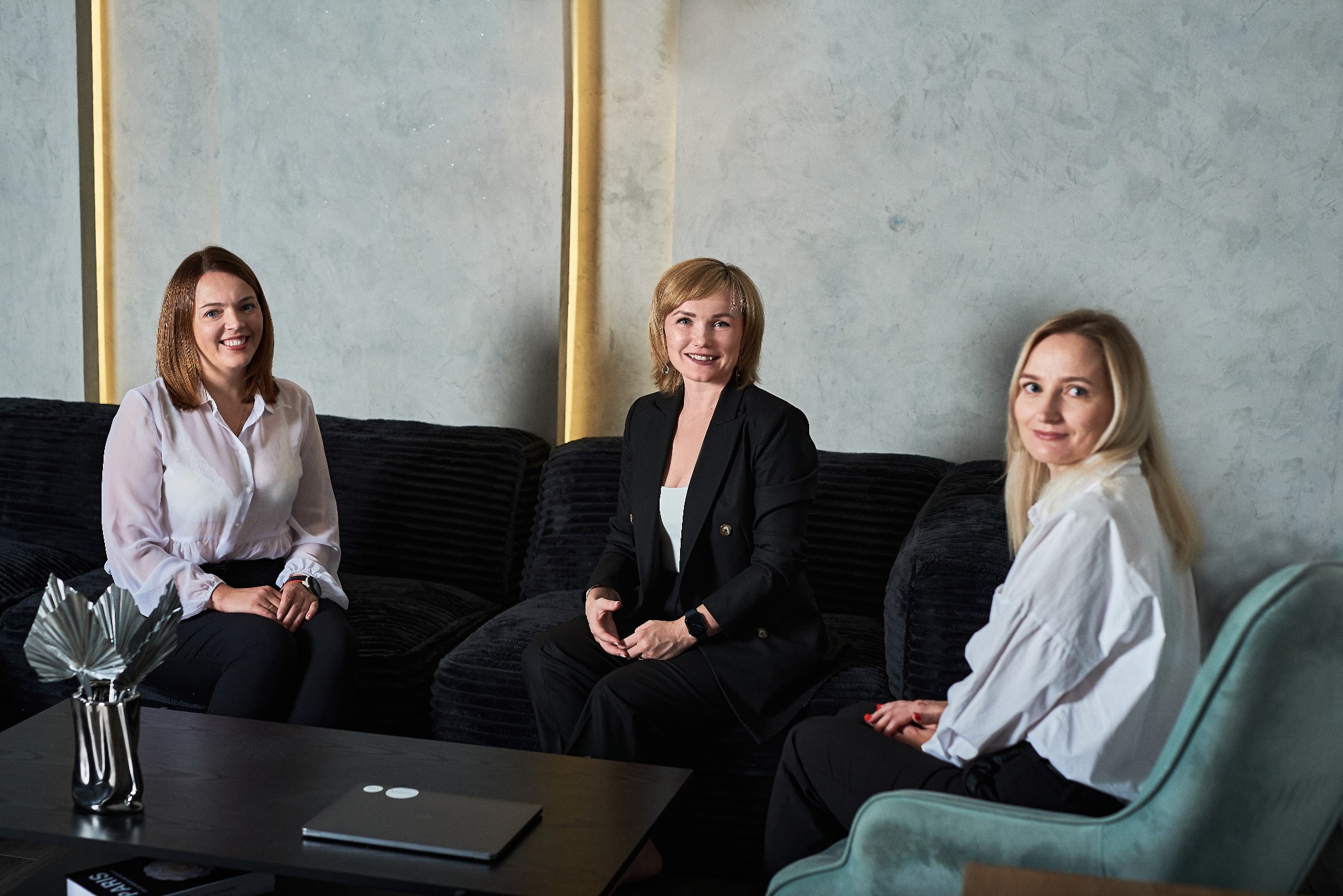 Trzy uśmiechnięte kobiety w eleganckich strojach siedzą na czarnej sofie w nowoczesnym biurze z szarą ścianą za nimi; na stoliku przed nimi srebrny wazon z suszonymi liśćmi i laptop.