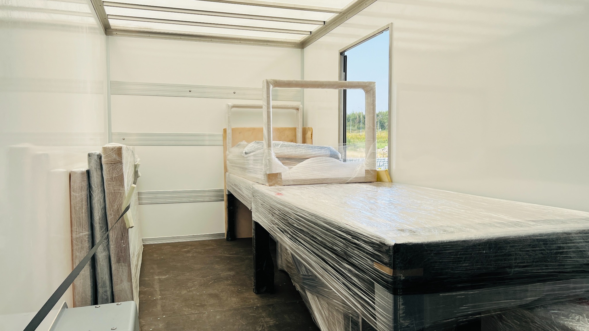 Wnętrze paki transportowej z meblami owiniętymi folią stretch, widoczne łóżko i elementy szafy, zabezpieczone do transportu.