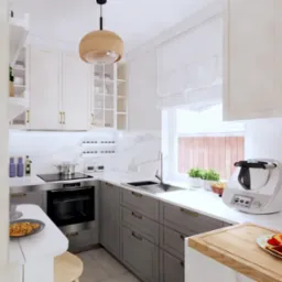 Jasna kuchnia w stylu klasycznym z szarymi szafkami, marmurowym blatem i nowoczesnym robotem kuchennym Thermomix obok okna z roletą rzymską.