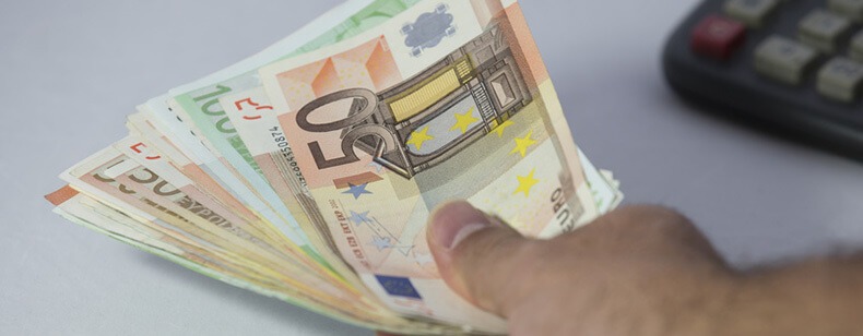 Dłoń trzymająca plik banknotów euro (100 i 50) na tle kalkulatora.