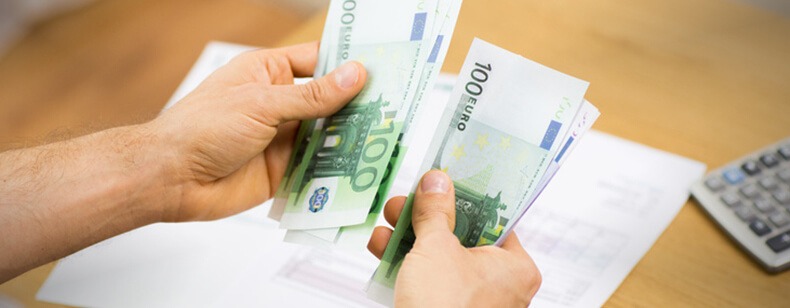 Dłonie liczą plik banknotów 100 euro, w tle kalkulator i dokumenty na drewnianym biurku.