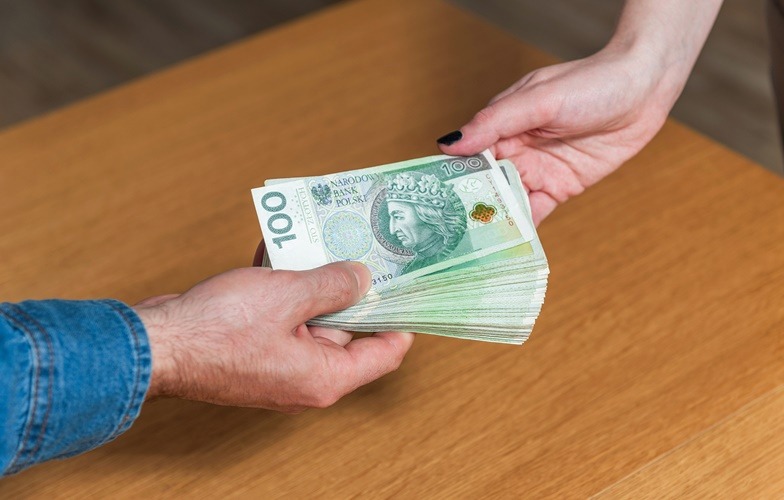 Przekazywanie pliku banknotów 100 zł między dwiema osobami na drewnianym stole.