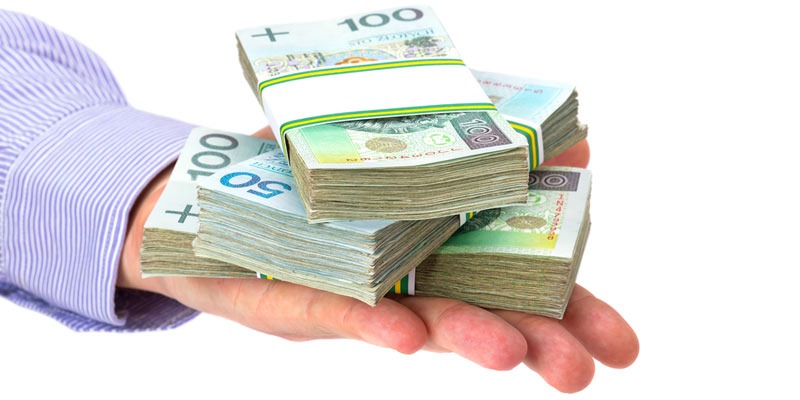 Dłoń w koszuli w paski trzymająca kilka plikow polskich banknotów o nominałach 50 i 100 złotych, spiętych gumkami.