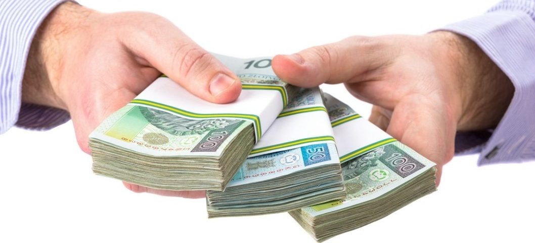 Dłonie w mankietach koszuli prezentują trzy spięte pakiety banknotów 100 i 50 złotowych na białym tle.