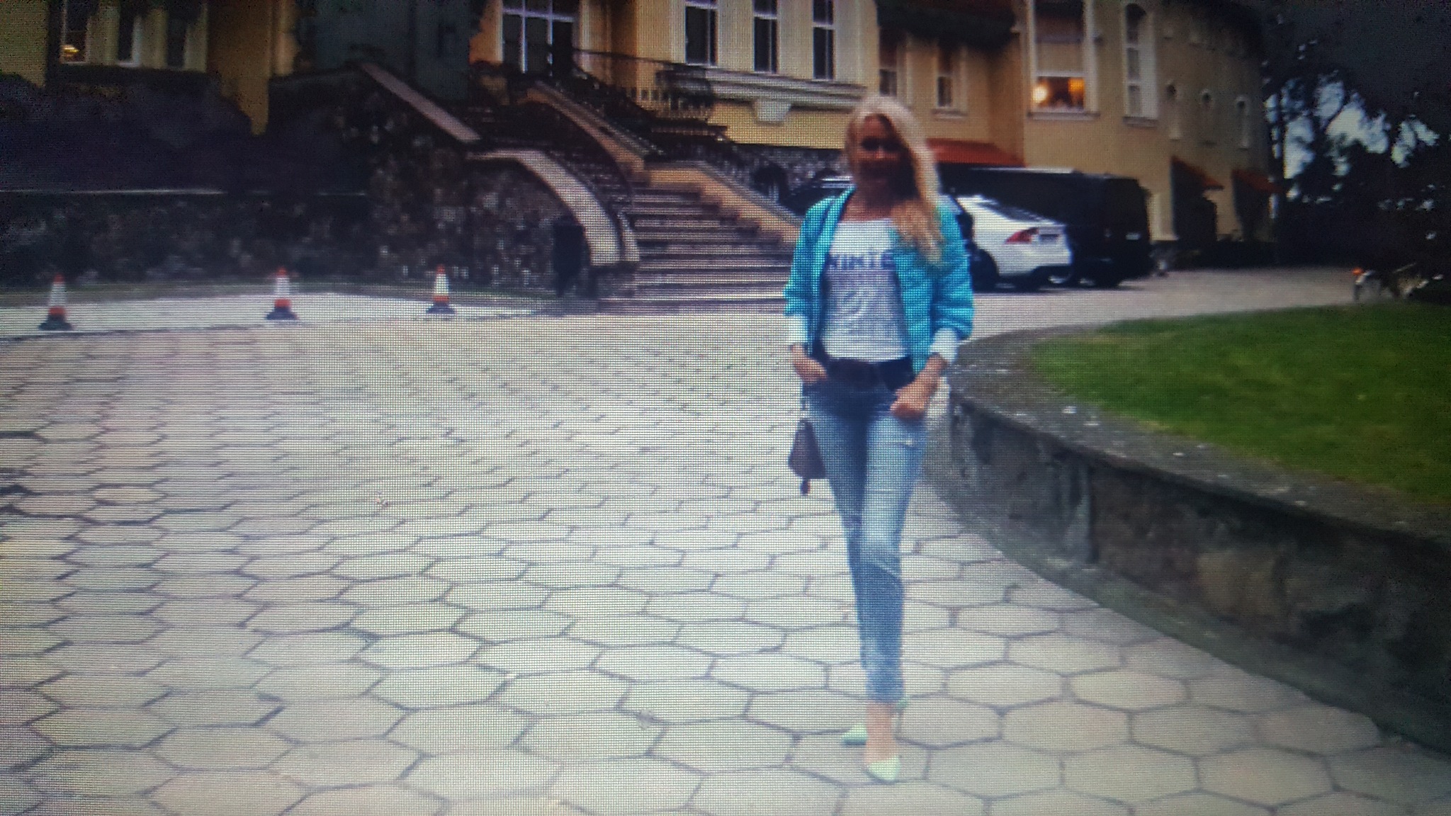 Kobieta w jeansowej kurtce i dżinsach stoi na chodniku z sześciokątną kostką brukową przed budynkiem z białymi schodami i pomarańczowymi pachołkami.