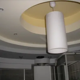 Wielopoziomowy sufit z oświetleniem punktowym i cylindryczną lampą wiszącą w łazience z szarą glazurą i widocznym fragmentem instalacji wentylacyjnej.