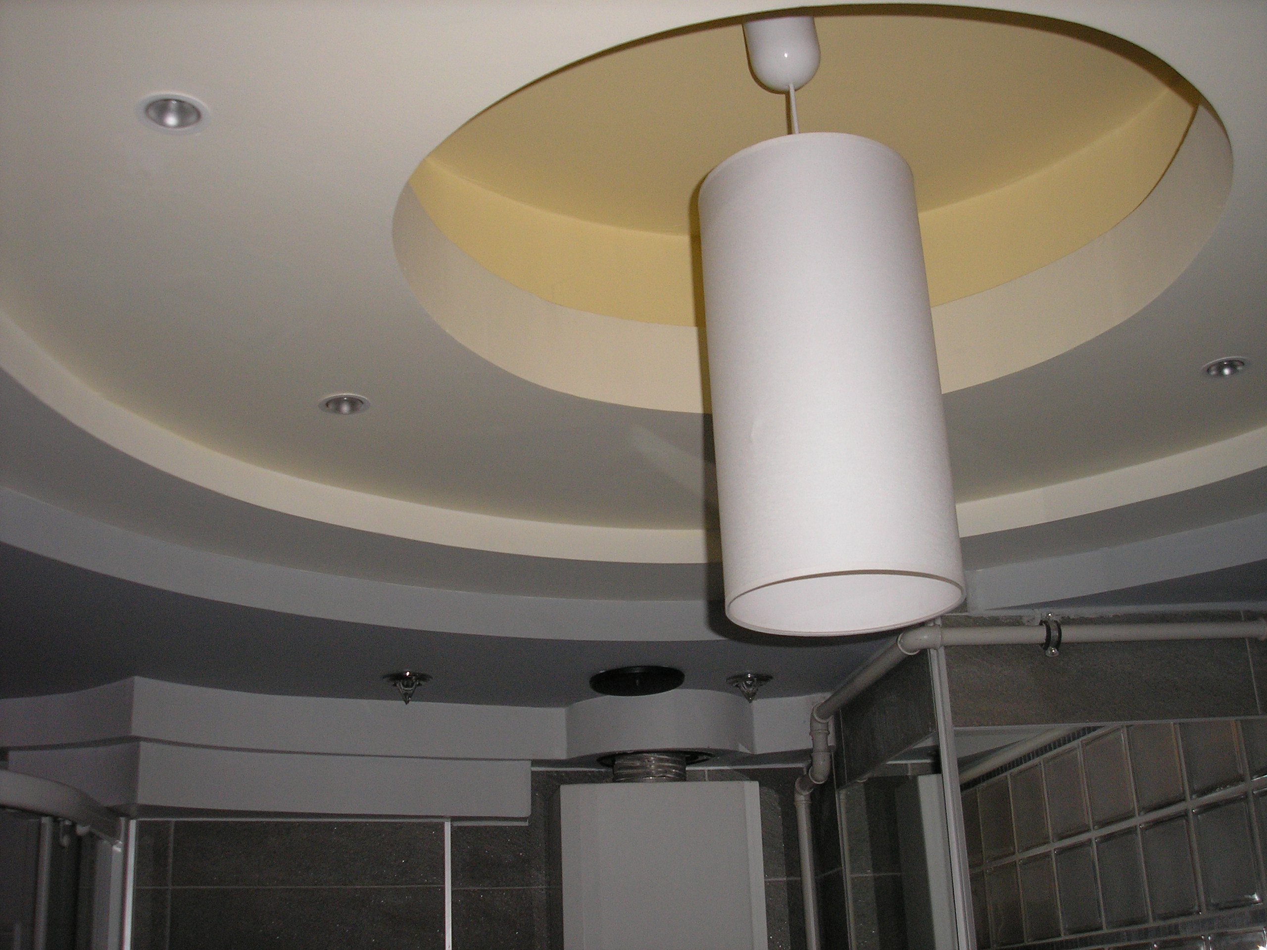 Wielopoziomowy sufit z oświetleniem punktowym i cylindryczną lampą wiszącą w łazience z szarą glazurą i widocznym fragmentem instalacji wentylacyjnej.