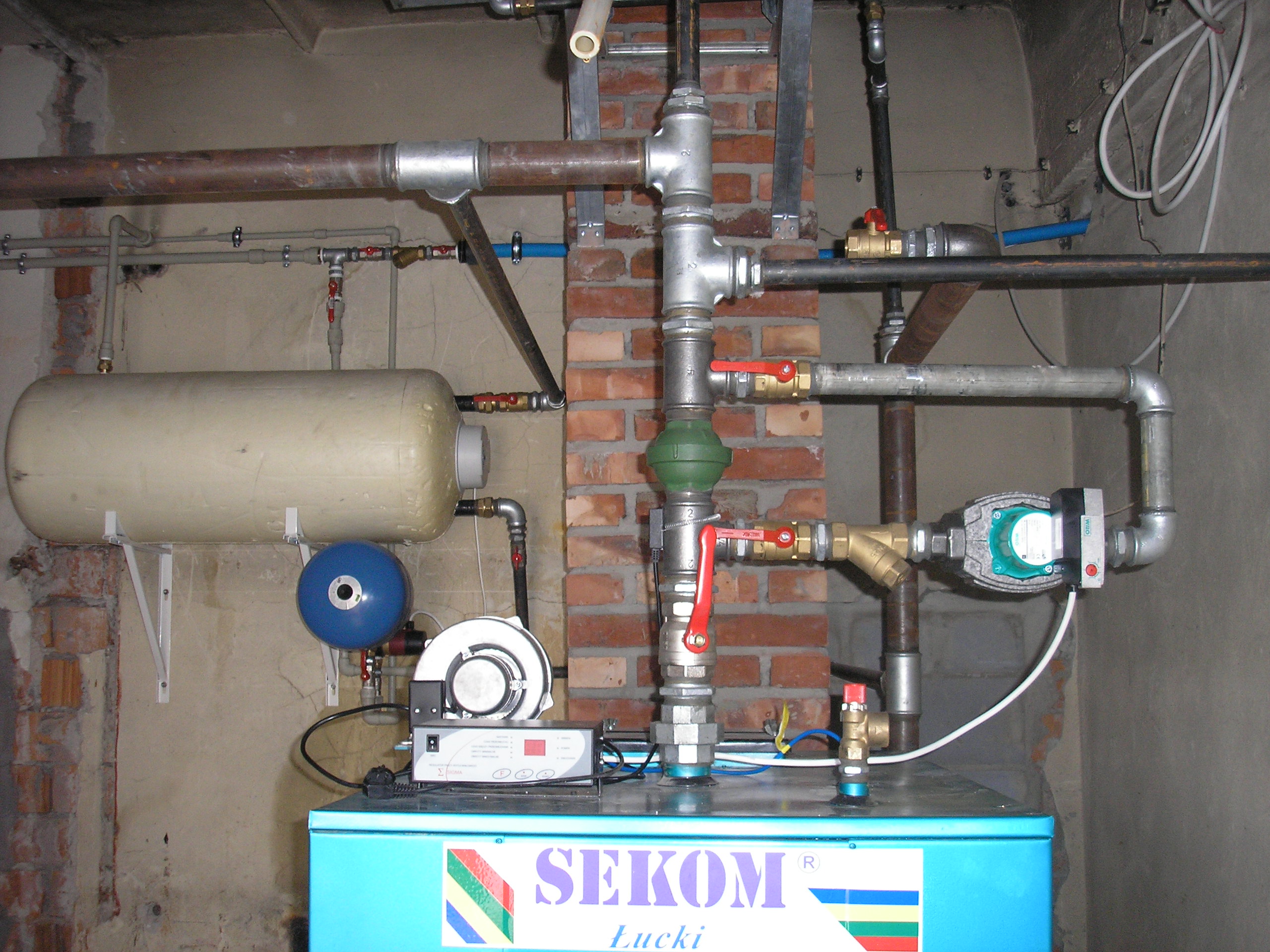 Kompleksowy system rur grzewczych, zaworów, zbiornika wyrównawczego i pompy obiegowej w piwnicy, z widocznym kotłem Sekom Łucki.