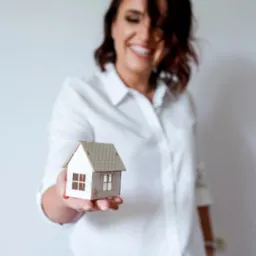 Uśmiechnięta kobieta w Częstochowie prezentuje miniaturowy model domu z drewna na dłoni, na jasnym tle.