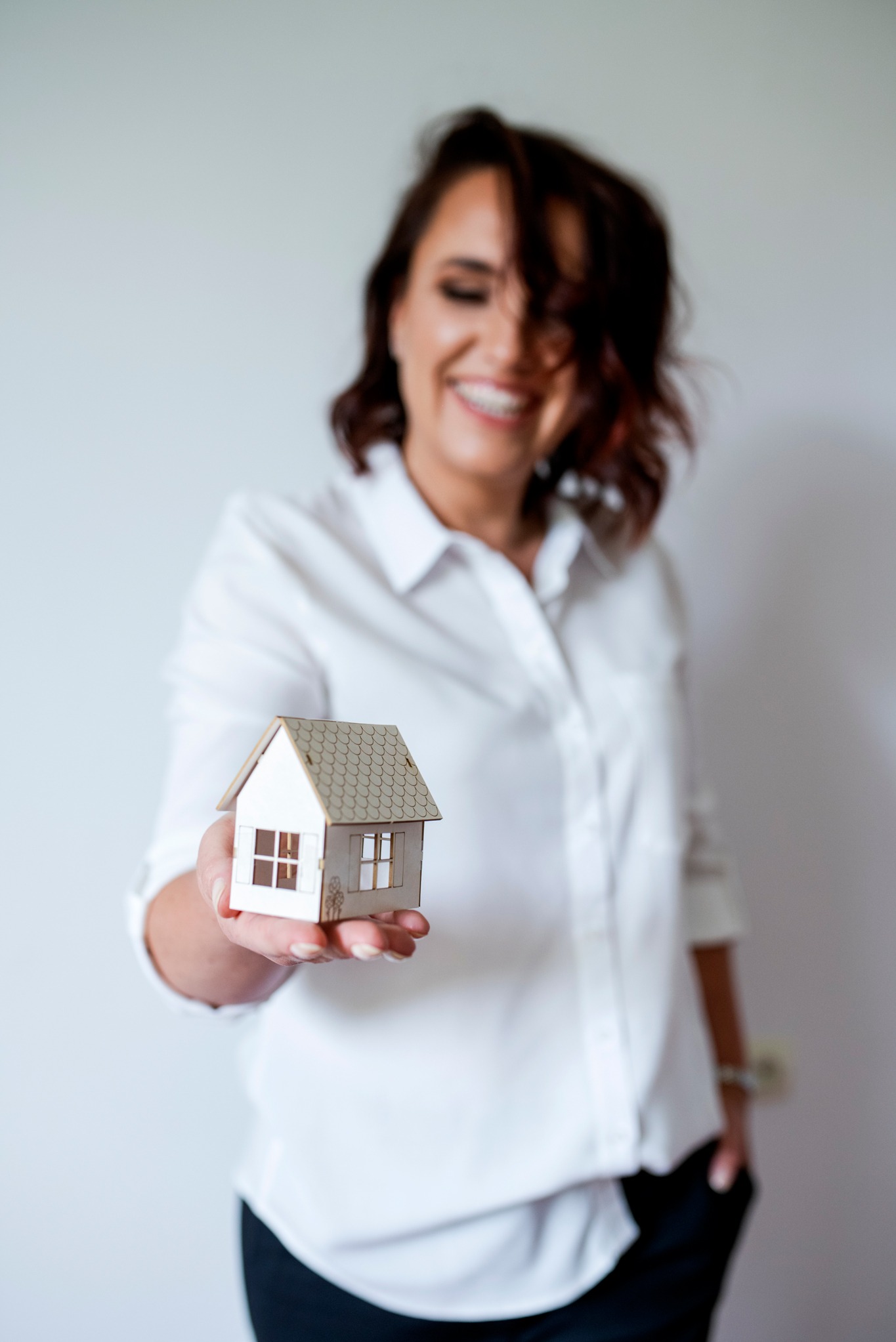 Uśmiechnięta kobieta w Częstochowie prezentuje miniaturowy model domu z drewna na dłoni, na jasnym tle.