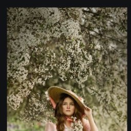 Jagoda Trzyna Photography - Portret młodej kobiety w słomkowym kapeluszu i różowej sukience, siedzącej w kwitnącym sadzie, trzymającej bukiecik białych kwiatów, z delikatnym oświetleniem i miękkim tłem.