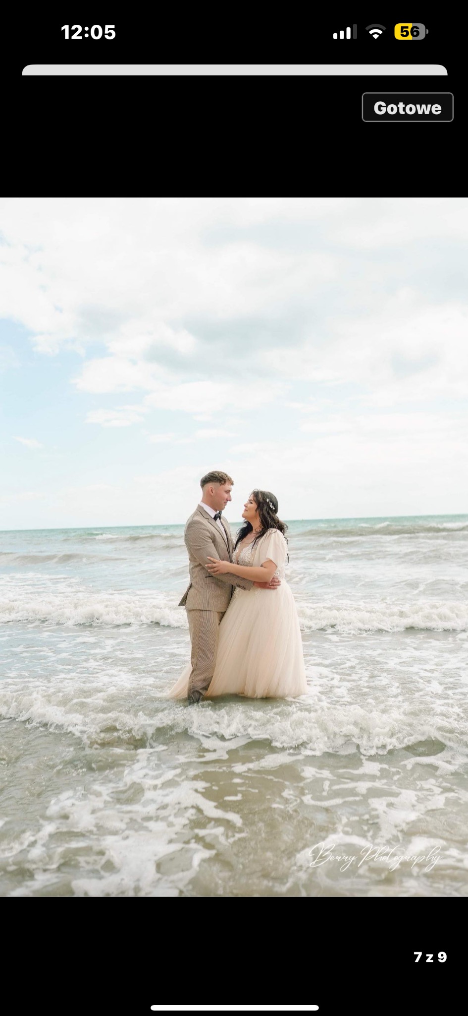 Para młoda w objęciach, stojąca w płytkiej wodzie morskiej; panna młoda w jasnej sukni, pan młody w brązowym garniturze w kratę, oboje patrzą na siebie z uśmiechem.