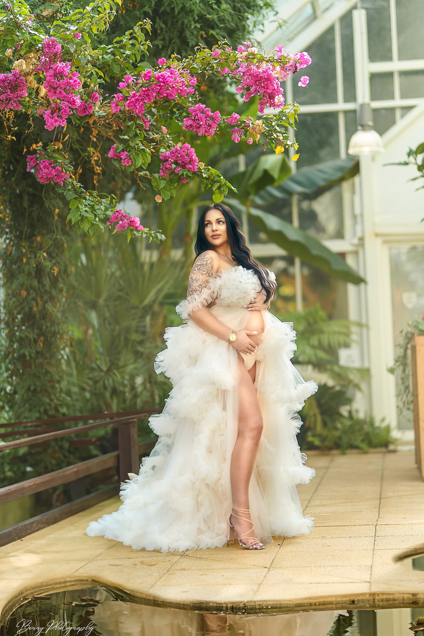 Kobieta w ciąży w tiulowej sukni, pozująca w oranżerii z kwitnącą bugenwillą i małym oczkiem wodnym.