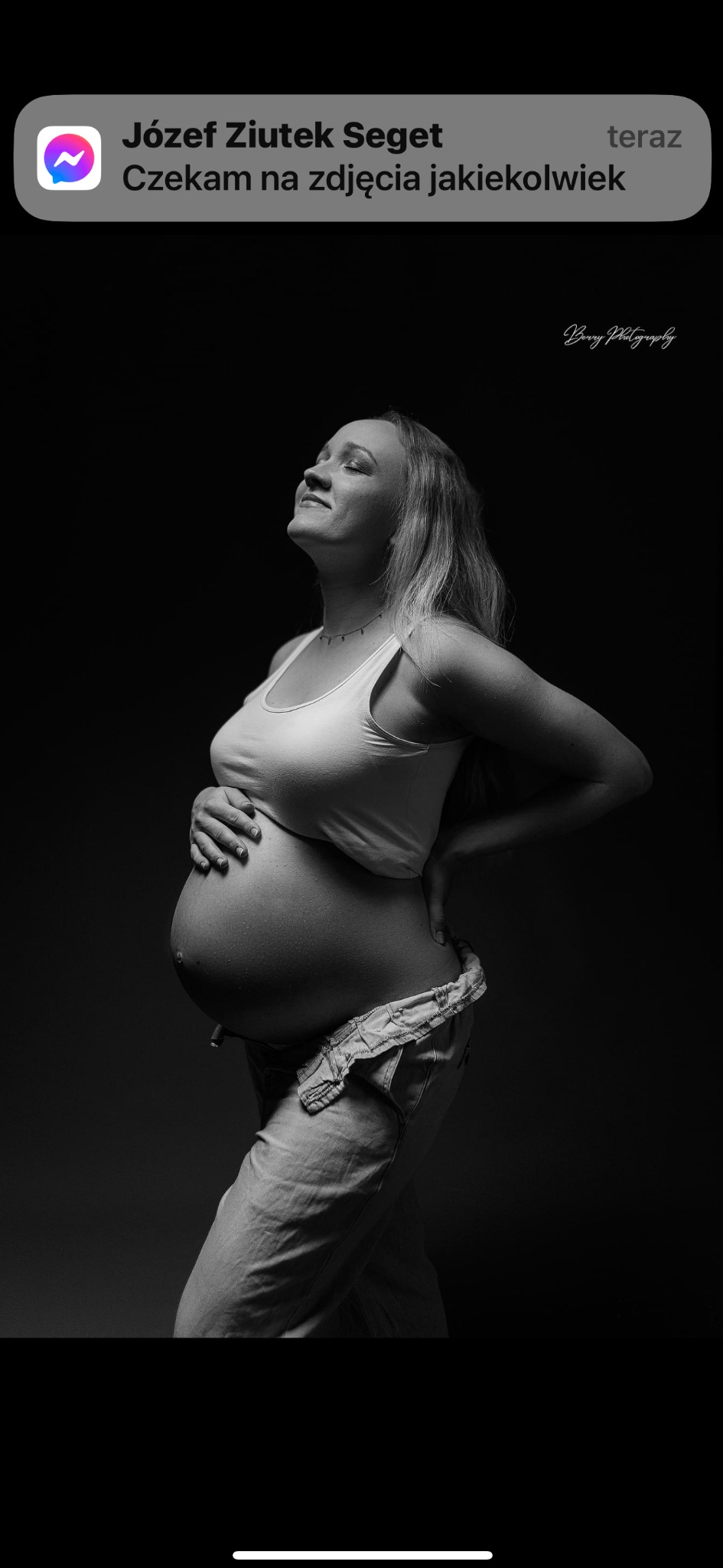 Czarno-białe zdjęcie kobiety w ciąży, w białym topie i rozpiętych dżinsach, z rękami na brzuchu i biodrze, głowa odchylona do tyłu, oczy zamknięte, na ciemnym tle.