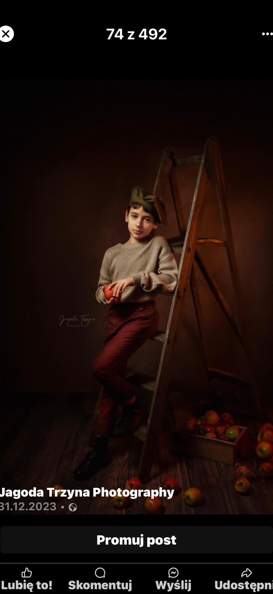 Portret chłopca w zielonej czapce, opartego o drewnianą drabinę, trzymającego czerwone jabłko na tle ciemnego studia z skrzynką pełną jabłek w dolnym rogu.