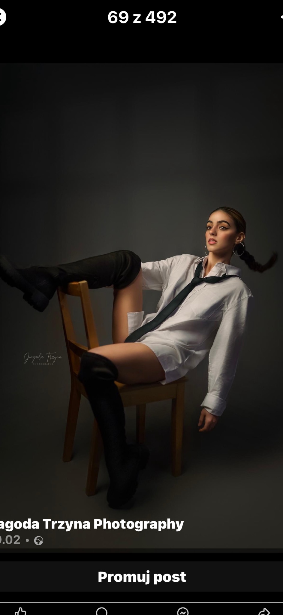 Portret kobiety w białej koszuli, czarnym krawacie i wysokich butach, pozującej na drewnianym krześle na szarym tle w studiu.