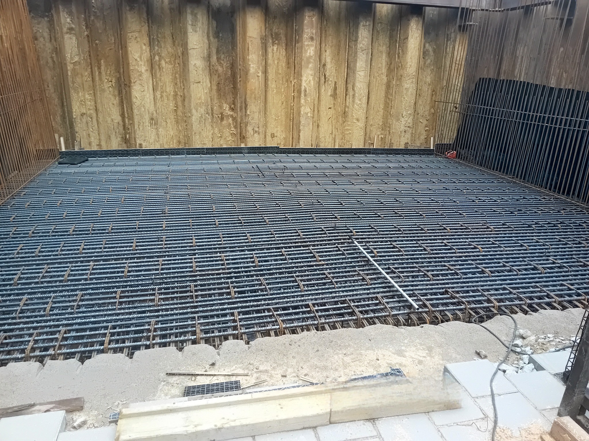 Siatka z prętów na fundamencie, przygotowana do zalania betonem. Widoczne drewniane szalunki i pionowe pręty. Prace ziemne na budowie w toku.