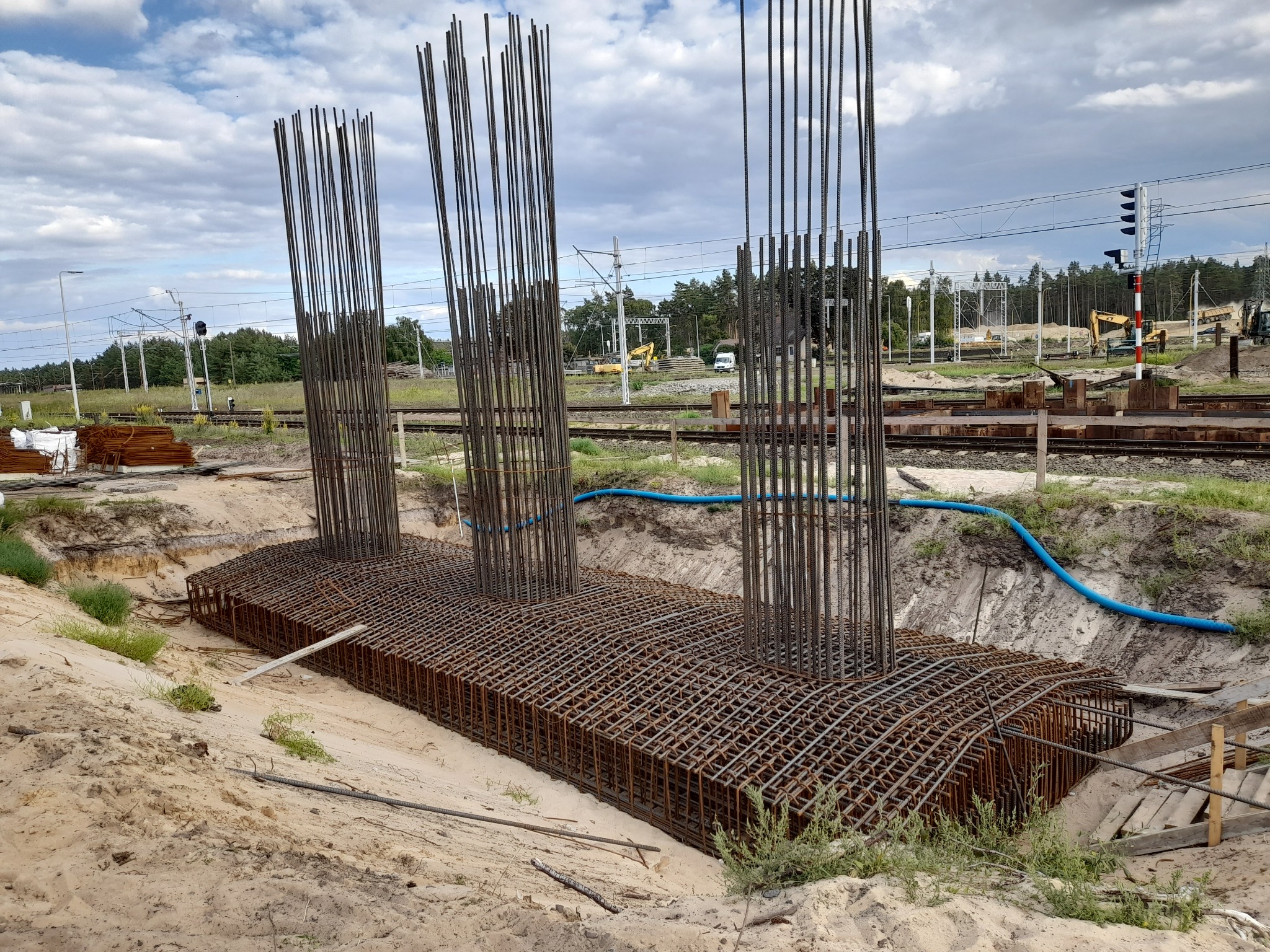 Zbrojenie fundamentu pod słupy na tle torów kolejowych i błękitnego nieba. Widoczne pionowe pręty i pozioma siatka zbrojeniowa w wykopie.