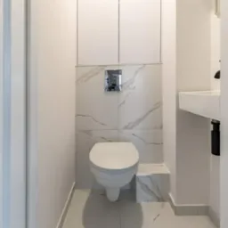 Kompaktowa toaleta w nowoczesnej łazience wykończonej płytkami imitującymi marmur, z zabudową nad stelażem i umywalką w rogu, widok z wejścia.