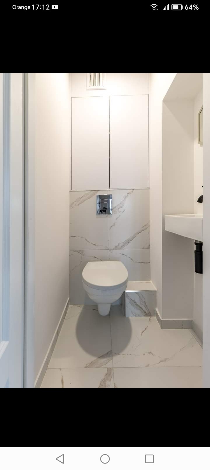 Nowoczesna toaleta podwieszana z białymi płytkami imitującymi marmur, zabudowa szafek nad nią i chromowany przycisk spłuczki, widok na fragment umywalki.