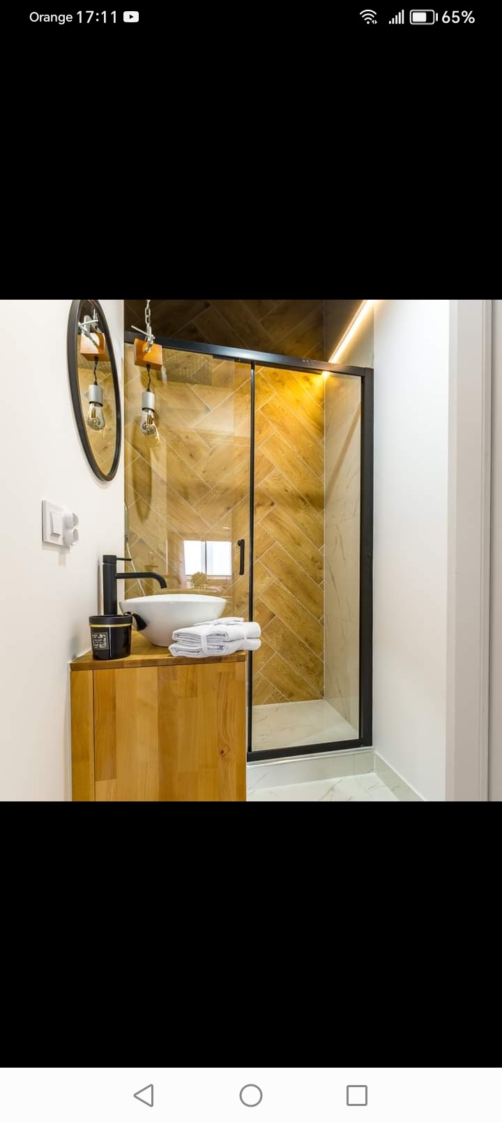 Nowoczesna łazienka z drewnianą szafką pod umywalką, czarną armaturą i kabiną prysznicową z drewnopodobnymi płytkami ułożonymi w jodełkę.