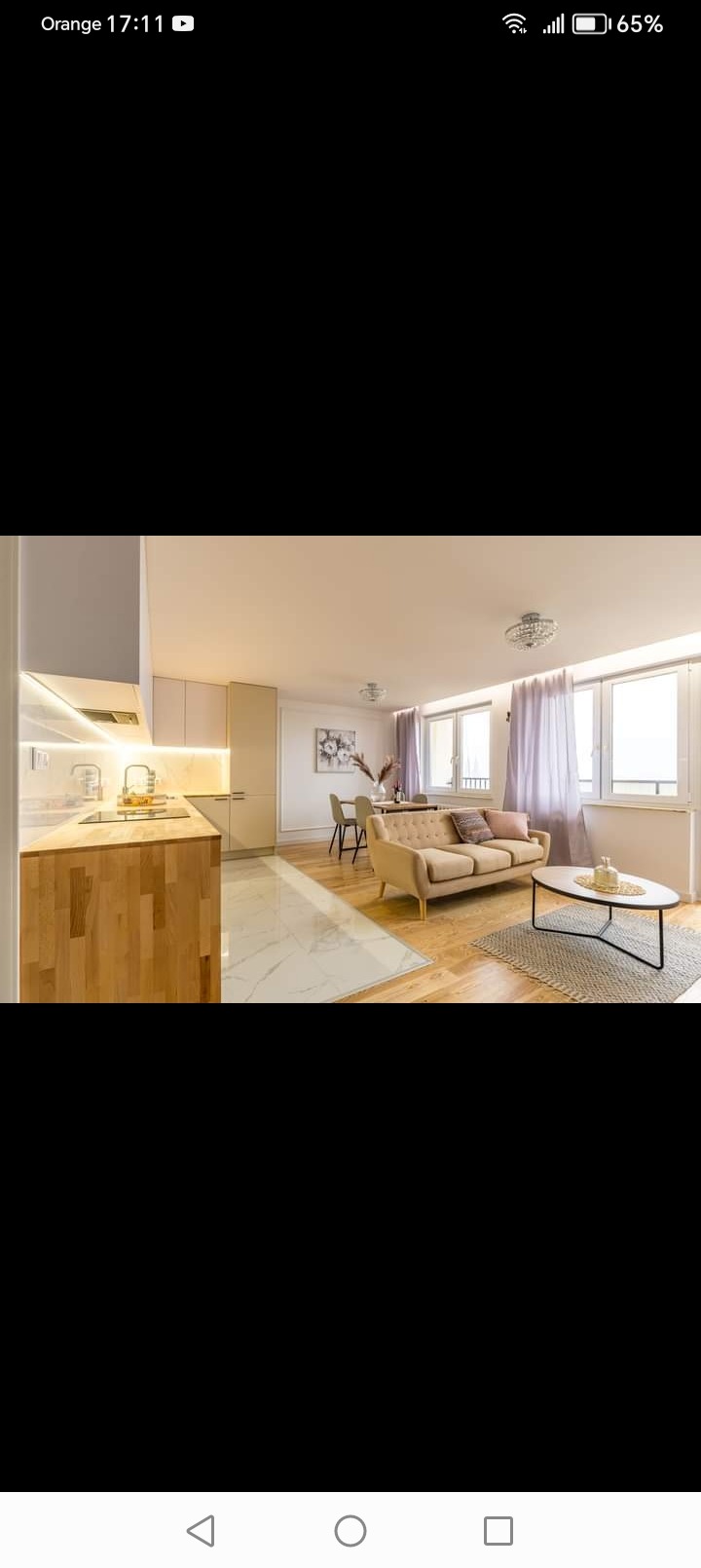 Jasne wnętrze po remoncie: marmurowe płytki podłogowe przechodzą w drewnianą podłogę, beżowa sofa z poduszkami, stół z krzesłami w tle, kuchnia z drewnianym blatem i białymi szafkami, okna z firanami.