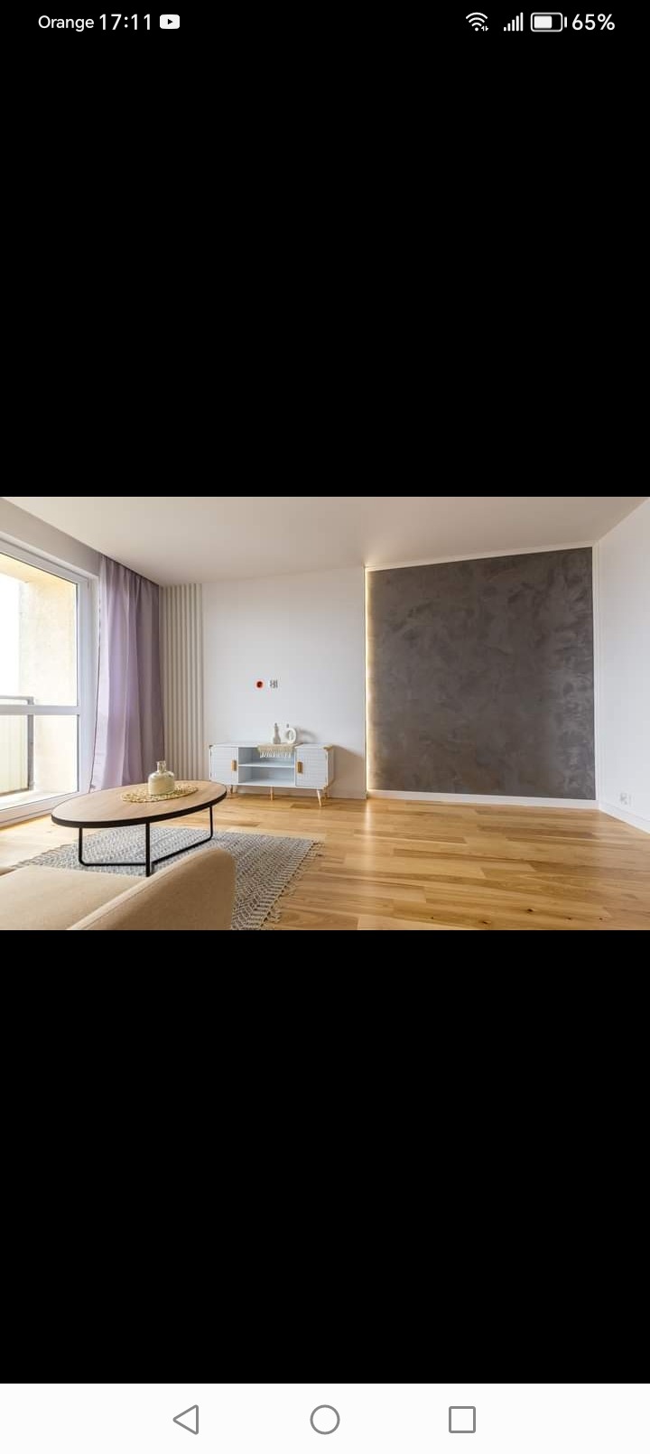 Jasne wnętrze salonu z drewnianą podłogą, szarą ścianą z efektem betonu, białym kredensem i okrągłym stolikiem kawowym na wzorzystym dywanie.