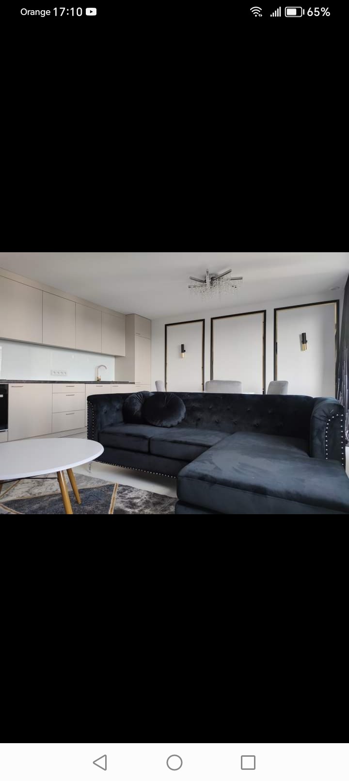 Nowoczesne wnętrze z aneksem kuchennym, ciemnogranatową sofą narożną z ozdobnymi ćwiekami i okrągłym stolikiem kawowym na wzorzystym dywanie.