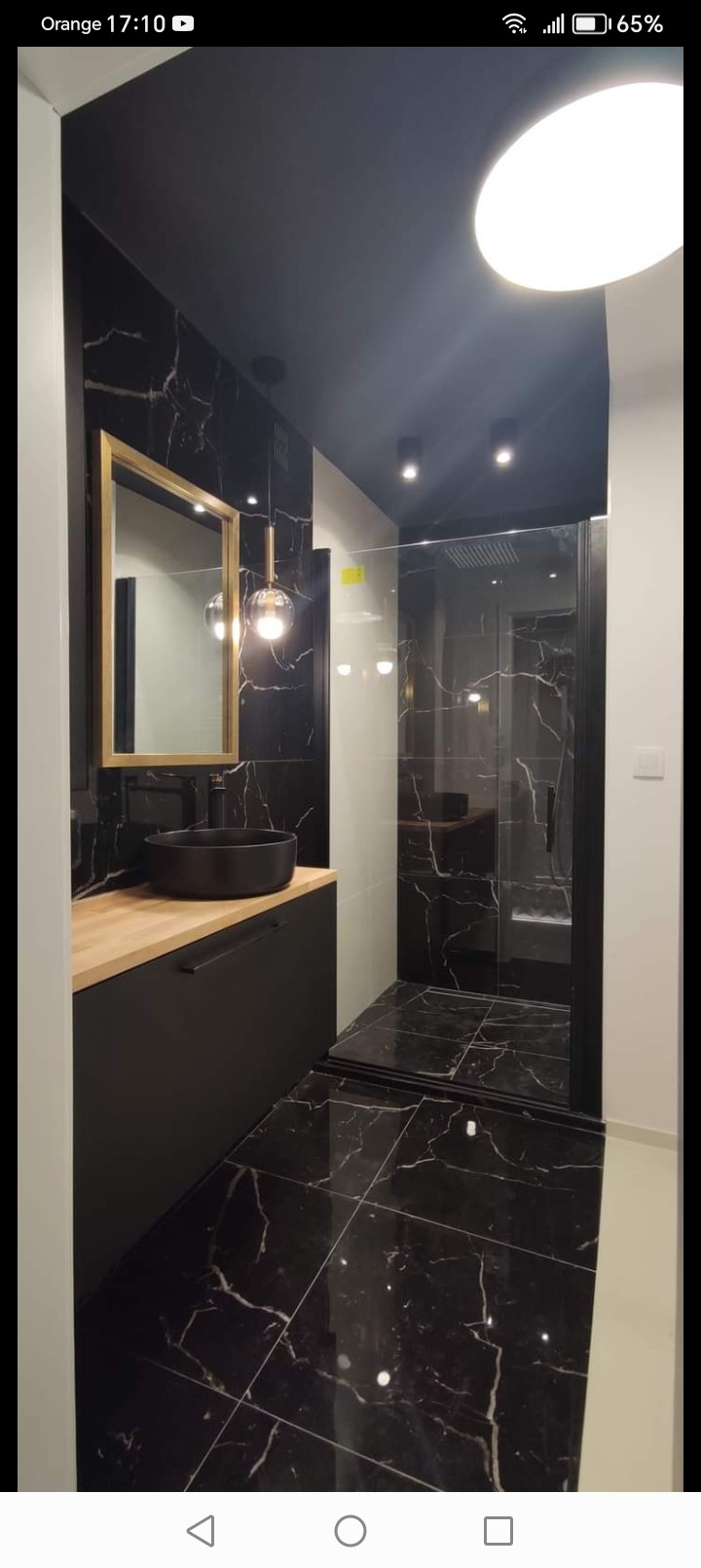 Nowoczesna łazienka w czerni i bieli z elementami drewna: czarne płytki imitujące marmur, złota rama lustra, okrągła umywalka nablatowa i kabina prysznicowa typu walk-in.