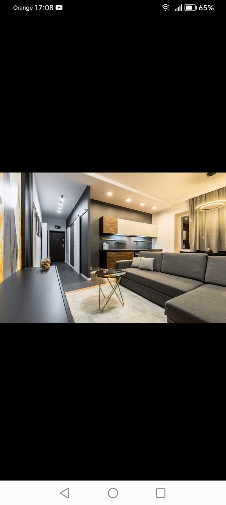 Nowoczesne wnętrze mieszkania z szarą sofą narożną, złotym stolikiem kawowym i minimalistyczną kuchnią w tle. Na ścianie dekoracyjna okładzina z motywem marmuru.