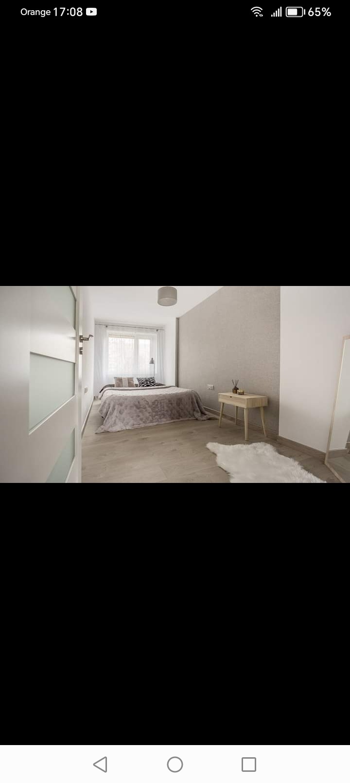 Jasna sypialnia z łóżkiem, drewnianą podłogą i puszystym dywanikiem, widok zza częściowo przeszklonych drzwi.