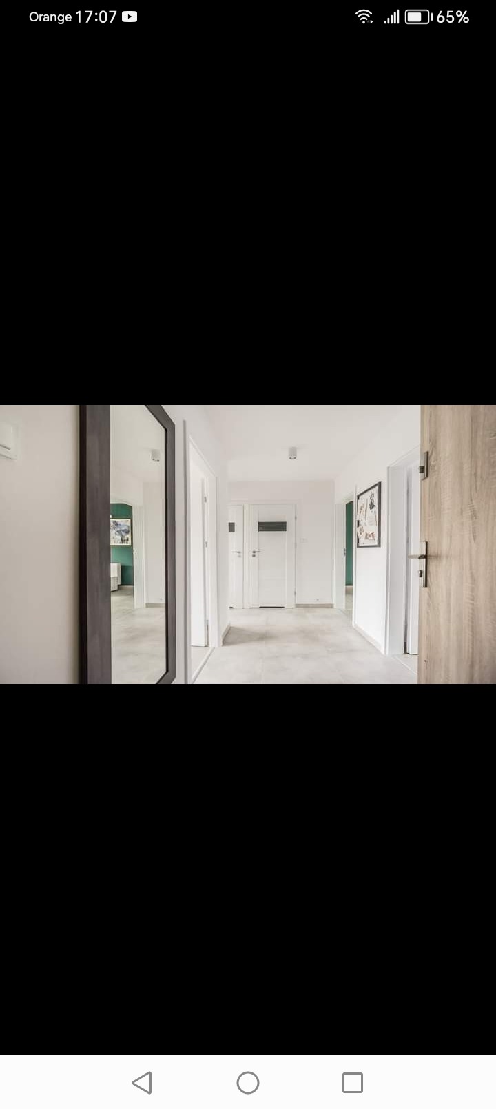 Nowoczesny korytarz z szarą podłogą, białymi ścianami i drzwiami, z widocznym lustrem w ciemnej ramie i elementami dekoracyjnymi.