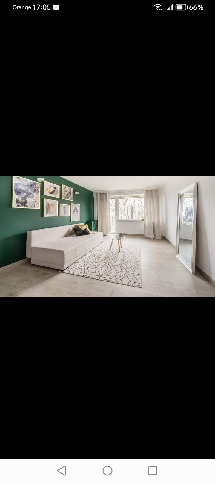 Jasny salon z zieloną ścianą, galerią obrazów, beżową sofą, geometrycznym dywanem i dużym lustrem opartym o ścianę.