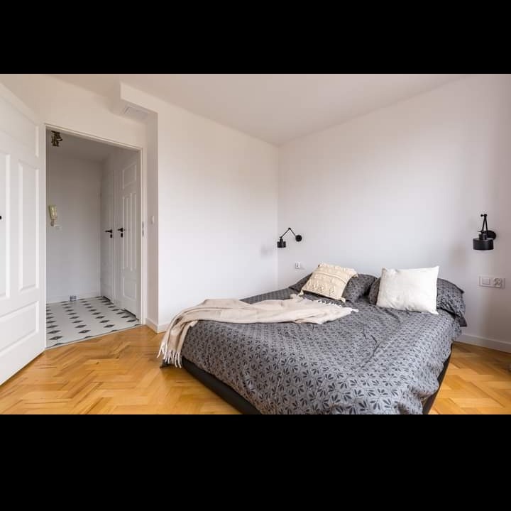 Jasna sypialnia z parkietem w jodełkę, szarą pościelą w drobny wzór i czarnymi kinkietami, widok na drzwi do łazienki z biało-czarną posadzką.