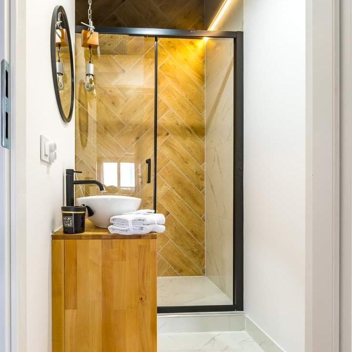 Nowoczesna łazienka z kabiną prysznicową wyłożoną płytkami imitującymi drewno w jodełkę, umywalką nablatową na drewnianej szafce i okrągłym lustrem podwieszonym na łańcuszku.