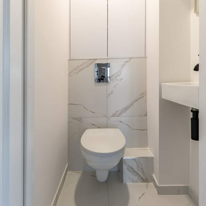Kompaktowa toaleta w nowoczesnej łazience wykończonej płytkami imitującymi marmur, z zabudową nad stelażem i umywalką w rogu, widok z wejścia.