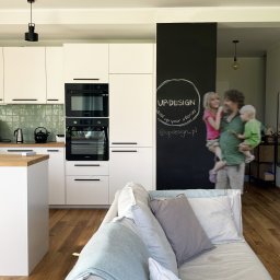 Anna Procner UP Design Interiors - Jasna kuchnia z zabudową, drewnianym blatem i zielonymi płytkami. Na ścianie czarna tablica z logo UP.DESIGN. W tle rodzina z dziećmi. Parkiet na podłodze.