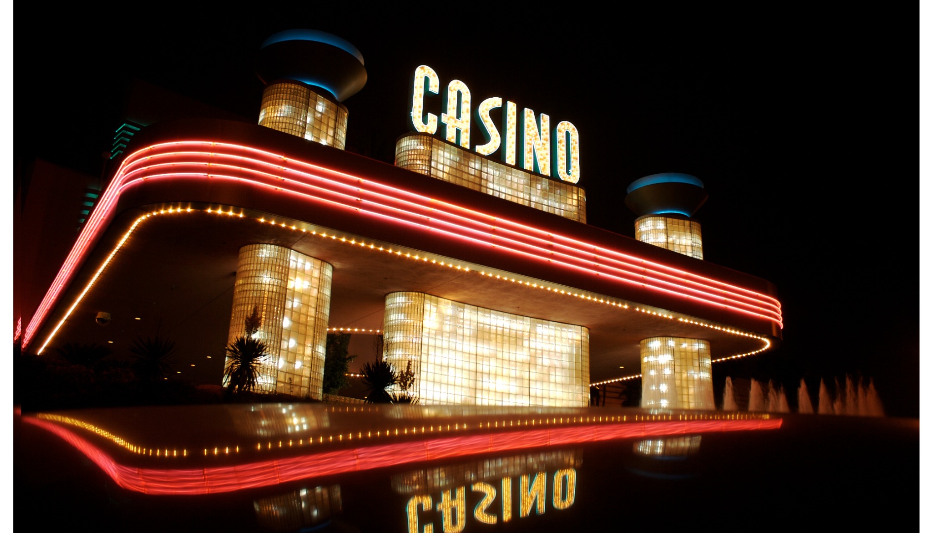 Fasada kasyna nocą z neonowym napisem 'CASINO' i podświetlonymi kolumnami z luksferów, odbijająca się w wodzie.