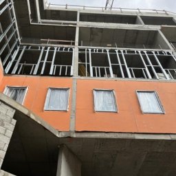 Global Invest development - Wznoszenie modułowego budynku wielopiętrowego: widok z dołu na pomarańczową elewację z oknami zabezpieczonymi folią, stalową konstrukcję szkieletową i betonowy szkielet budynku.