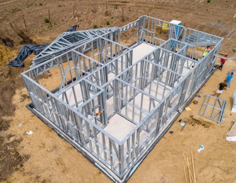 Widok z góry na stalowy szkielet domu w trakcie budowy, widoczni pracownicy w odblaskowych kamizelkach, elementy konstrukcyjne dachu leżące obok.