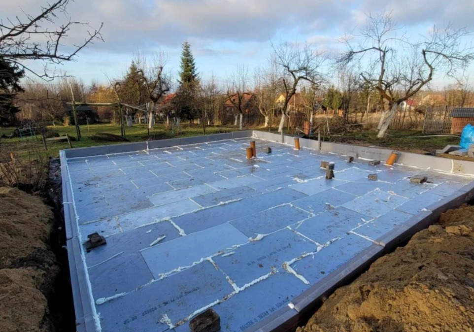 Przygotowany fundament pod budynek z izolacją z płyt styropianowych, uszczelniony pianką montażową, widoczne wystające rury kanalizacyjne i fragmenty cegieł, w tle ogród z drzewami.