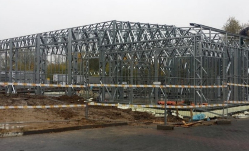 Stalowa konstrukcja szkieletowa domu w trakcie budowy, widoczne elementy mocujące, zabezpieczenia budowlane i częściowo ułożona izolacja fundamentów.