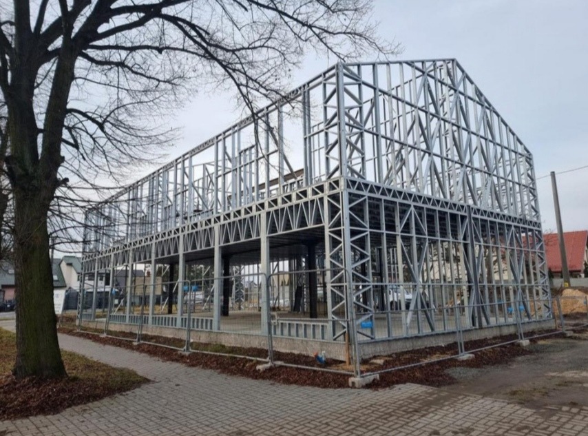 Konstrukcja szkieletowa dwukondygnacyjnego budynku w trakcie montażu, widoczna z zewnątrz na tle drzewa i zabudowań.