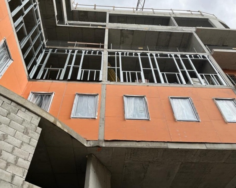 Wznoszenie modułowego budynku wielopiętrowego: widok z dołu na pomarańczową elewację z oknami zabezpieczonymi folią, stalową konstrukcję szkieletową i betonowy szkielet budynku.