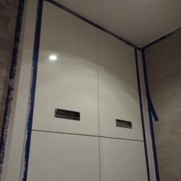 Usługi remontowo budowlane Adrian Matwiejczuk - Biała szafka łazienkowa z minimalistycznymi uchwytami, zabezpieczona niebieską taśmą malarską w trakcie prac wykończeniowych, widoczna w narożniku łazienki z oświetleniem sufitowym.