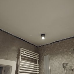 Usługi remontowo budowlane Adrian Matwiejczuk - Szary, matowy grzejnik drabinkowy w łazience z mozaikowymi płytkami, widoczna kabina prysznicowa z szarą ramą i okrągła lampa sufitowa