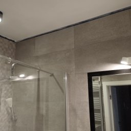 Usługi remontowo budowlane Adrian Matwiejczuk - Szklana kabina prysznicowa z chromowaną armaturą i deszczownicą, obłożona beżowymi płytkami w dużej formatce, widoczna fuga silikonowa na styku ściany i sufitu, odbicie wnętrza łazienki w lustrze.