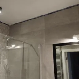 Szklana kabina prysznicowa z chromowaną armaturą i deszczownicą, obłożona beżowymi płytkami w dużej formatce, widoczna fuga silikonowa na styku ściany i sufitu, odbicie wnętrza łazienki w lustrze.