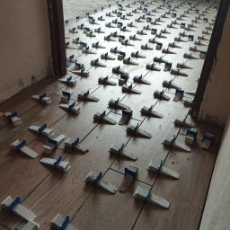 Usługi remontowo budowlane Adrian Matwiejczuk - Układanie płytek podłogowych z użyciem klipsów poziomujących. Widoczne brązowe płytki imitujące drewno i biało-niebieskie klipsy. Próg drzwiowy w tle.